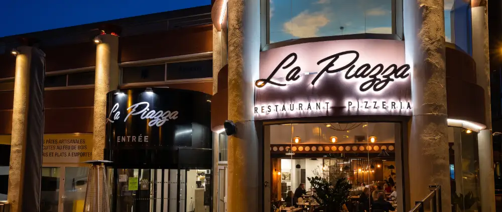 La façade de La Piazza de nuit : enseigne lumineuse « La Piazza, restaurant pizzeria » et entrée vitrée