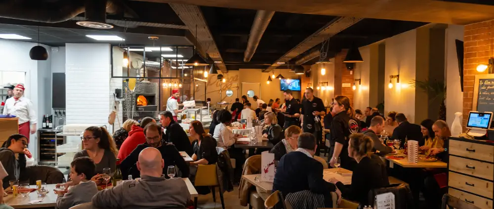 La salle pleine un soir de service, le pizzaïolo devant le four à bois
