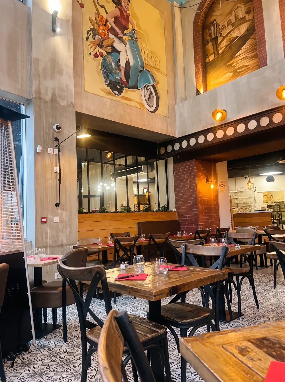 La salle de La Piazza : tables en bois, chaises bistrot et grande fresque d’une femme en Vespa
